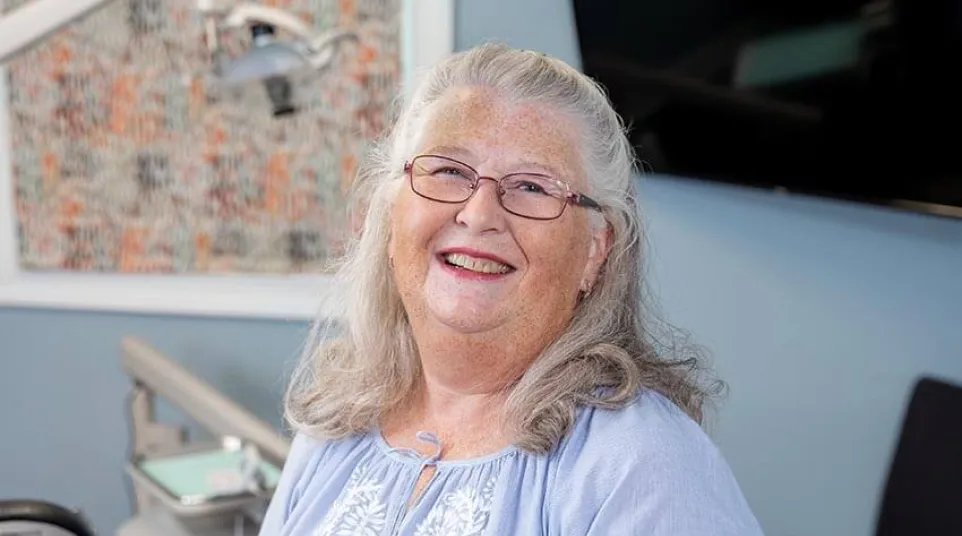 Smiling elderly woman with glasses and gray hair wearing a blue embroidered blouse in a bright room.