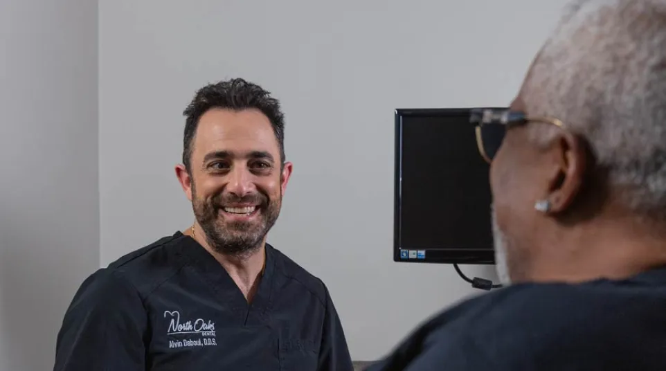 Dentist in black scrubs smiling and talking to an older male patient in a consultation room.