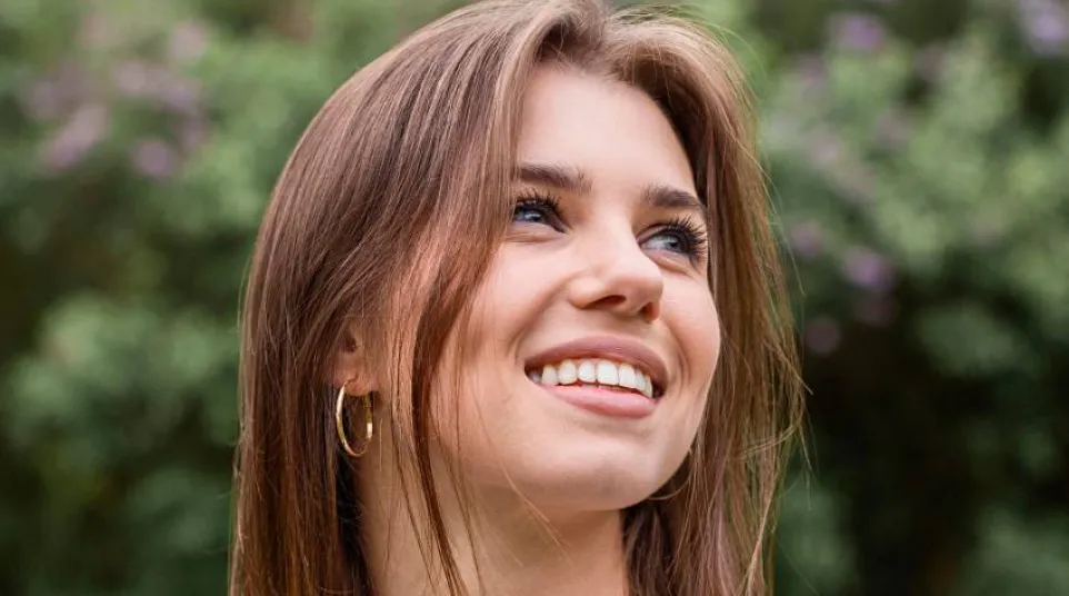 Smiling young woman with brown hair and hoop earrings outdoors with greenery and purple flowers behind her