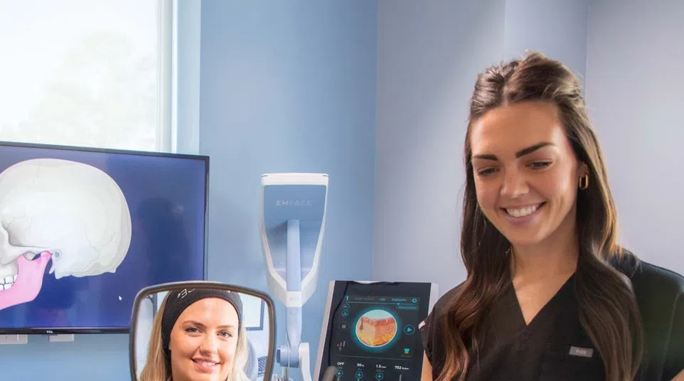 Dental patient looks in handheld mirror while dentist explains jaw anatomy with skull images on screen.