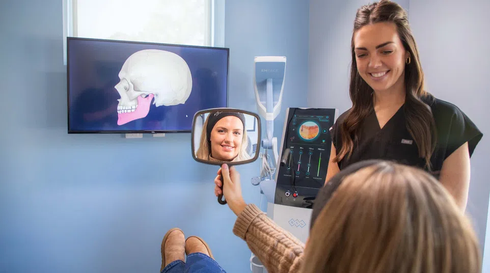Dental patient looks in handheld mirror while dentist explains jaw anatomy with skull images on screen.