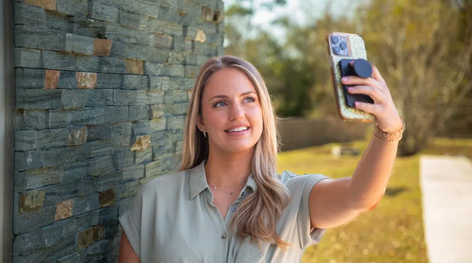 Young woman with blonde hair wearing a light shirt takes a selfie outdoors by a stone wall on a sunny day