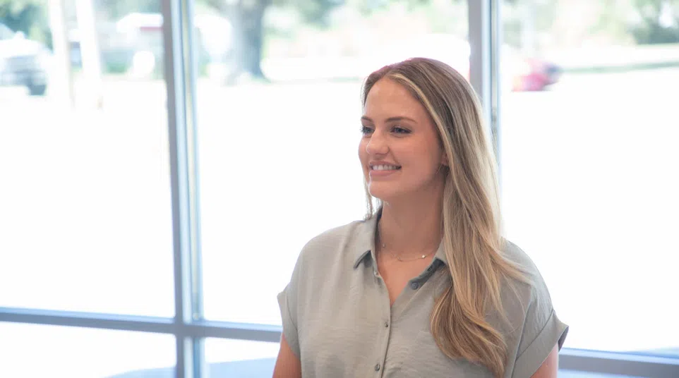 Smiling woman with long blonde hair wearing a grey blouse standing near large windows in bright room.
