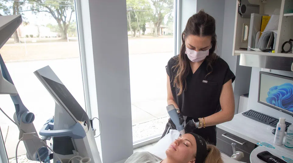 A woman receiving a facial treatment from a skincare professional wearing a mask in a modern clinic room.
