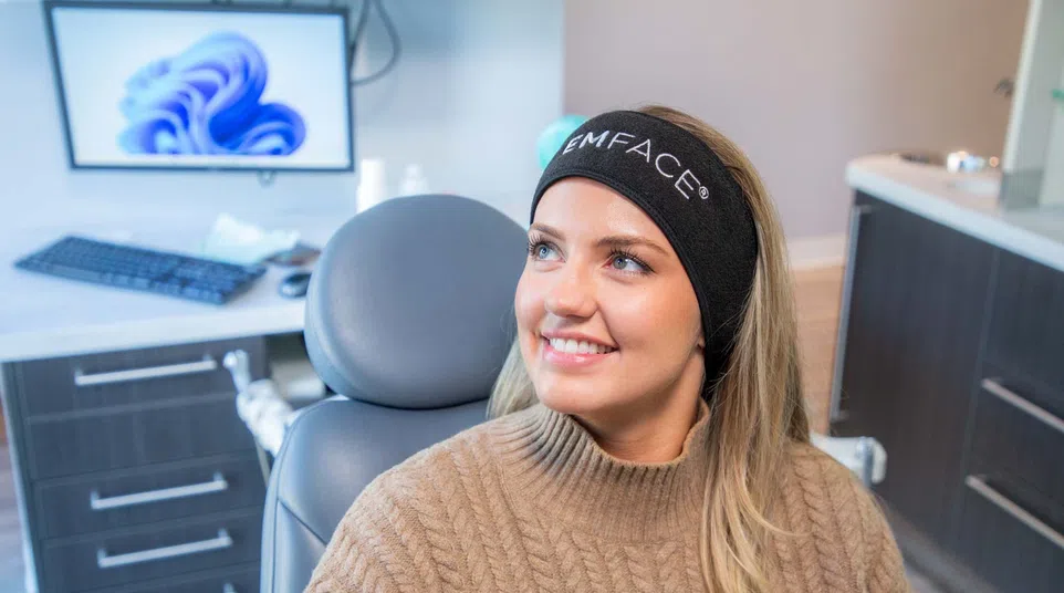 Smiling woman wearing a black EMFACE headband sitting in a modern clinic chair with computer in background