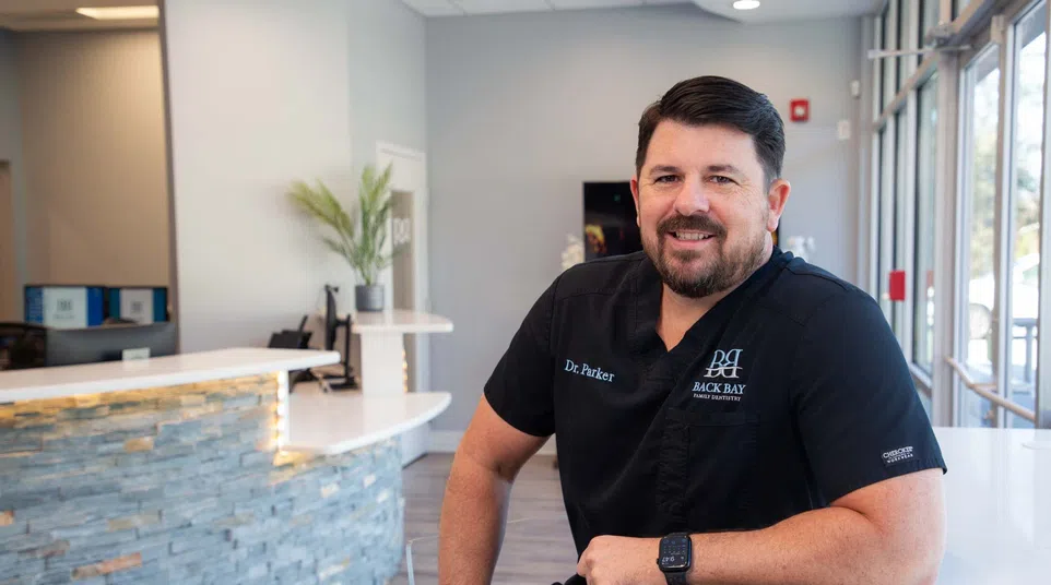 Smiling male doctor in black scrubs leans on white counter in modern medical office reception area