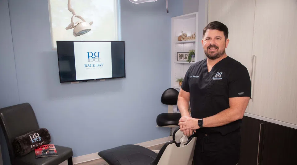 Dentist in black scrubs standing beside dental chair in modern clinic with Back Bay Dentistry logo on screen