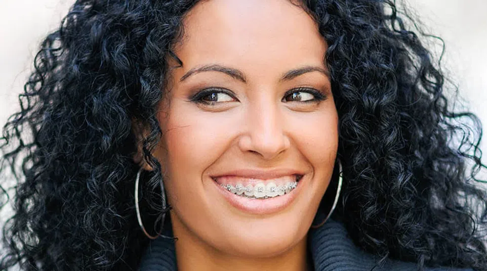 Smiling woman with curly black hair wearing braces and hoop earrings, dressed in a dark jacket outdoors.