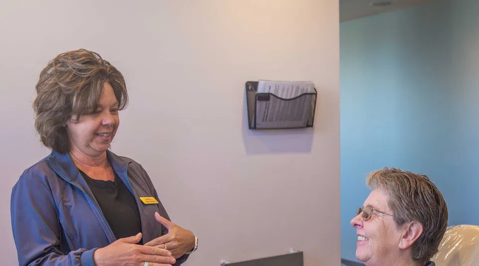 Dental hygienist talks to smiling patient sitting in a dental chair in a clean clinic room.