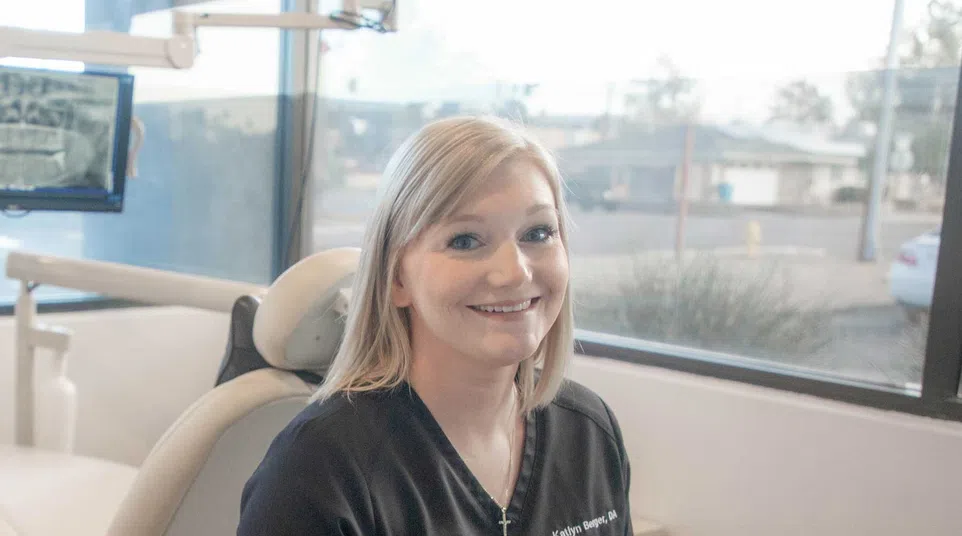 Smiling female dental professional sitting at a desk in a bright clinic with dental chair in background