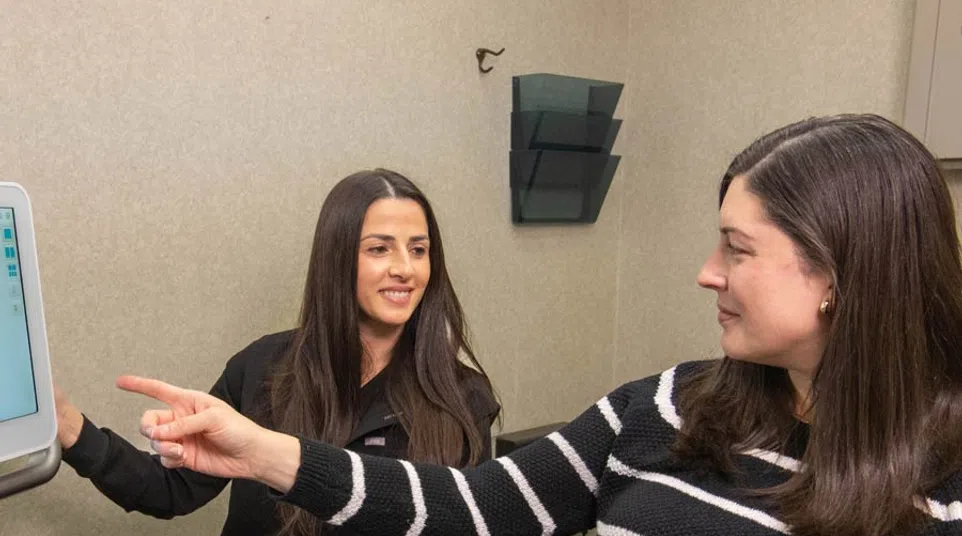Woman in striped sweater pointing at a screen while another woman in black observes and smiles in an office setting
