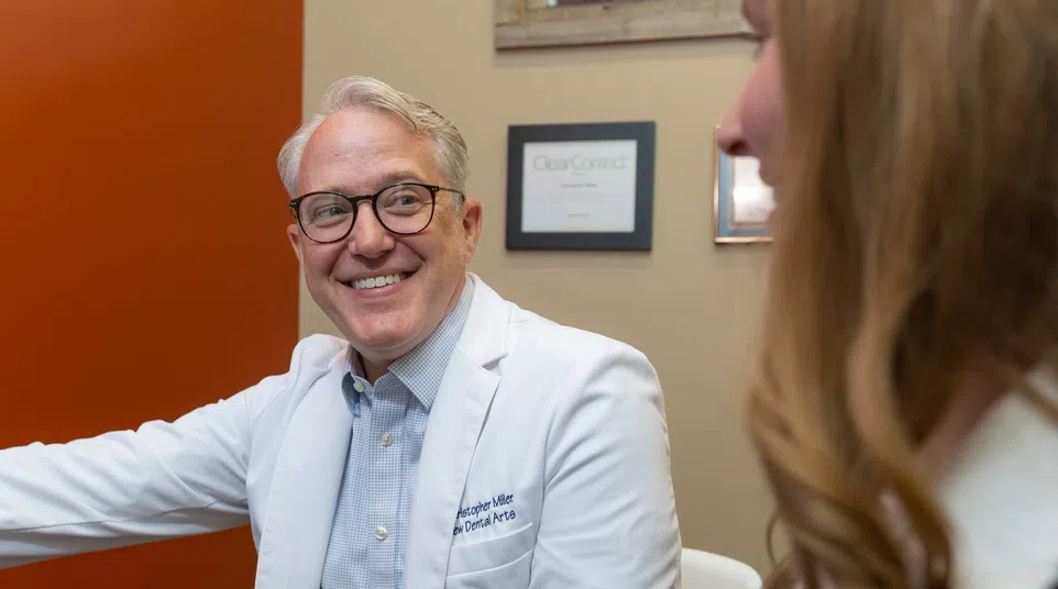 Smiling male dentist in white coat talking to a female patient in a dental office with certificates on the wall.