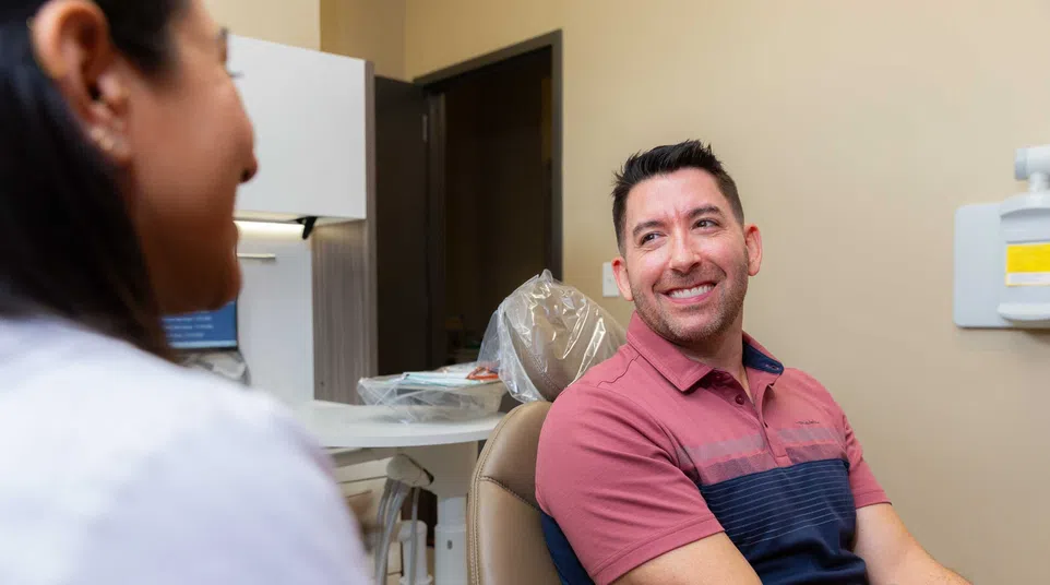 Man smiling while sitting in a dental chair talking to a dentist in a bright dental office.