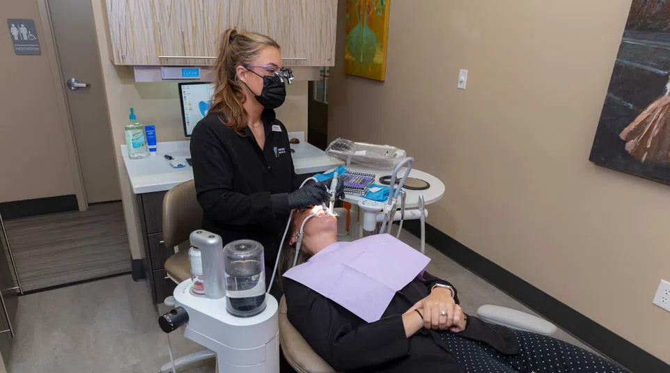Dentist wearing black mask treats patient lying back in dental chair during oral procedure in clinic.