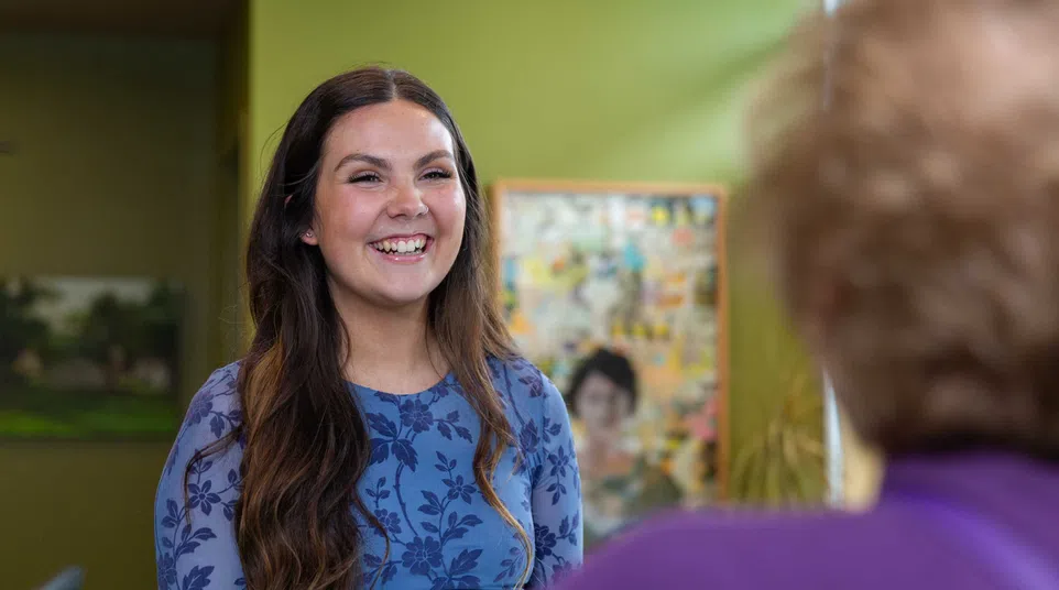 Smiling woman with long brown hair wearing a blue floral dress engaged in conversation indoors.
