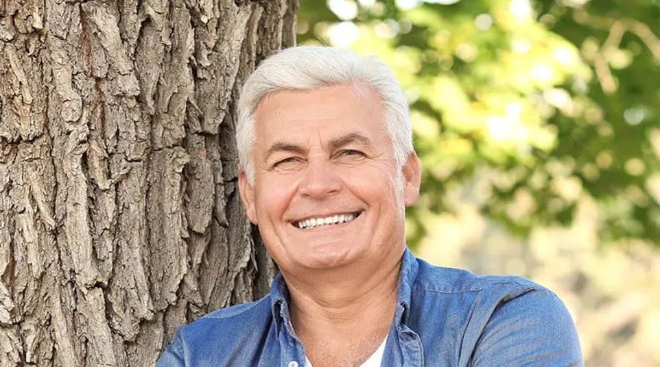 Smiling senior man in a denim shirt leaning against a tree, exuding warmth and confidence in natural light.