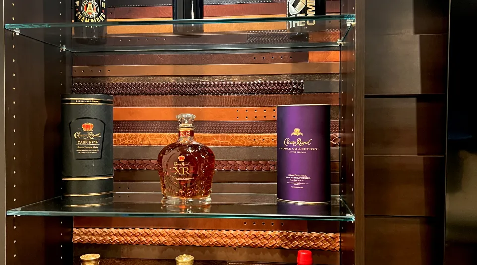 Close up of whisky bottles including Crown Royal and Maker's Mark bourbon on glass shelves with a wooden background in a whisky room