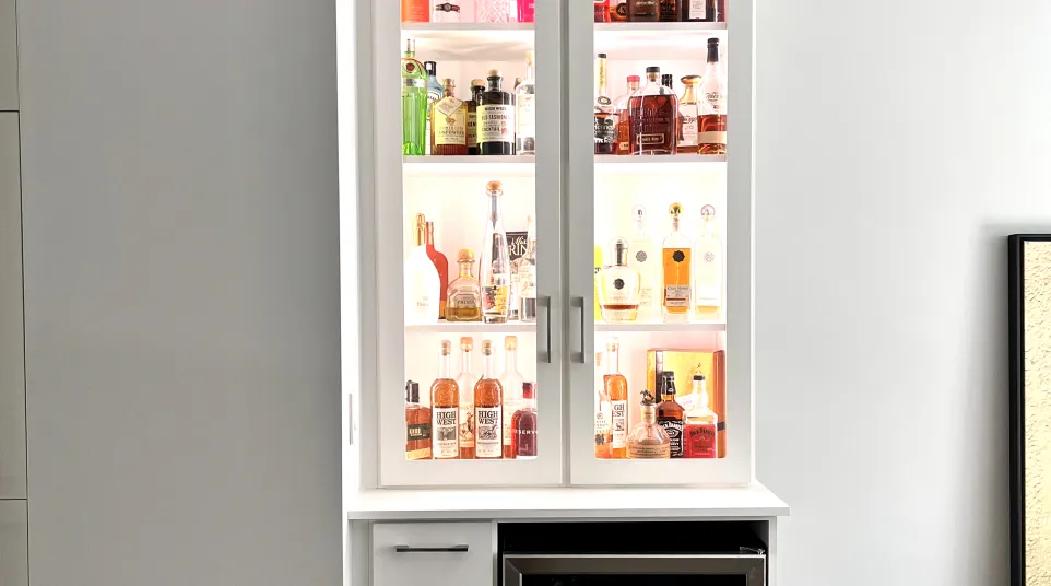 Home bar with liquor bottles displayed in a white cabinet with glass doors and accent lighting above a mini refrigerator