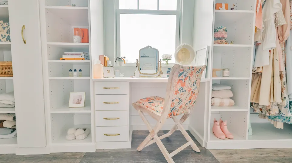 Walk in closet with white shelves, glass doors, white vanity area with floral cushioned chair, a mirror, hanging clothes in stylish dressing room
