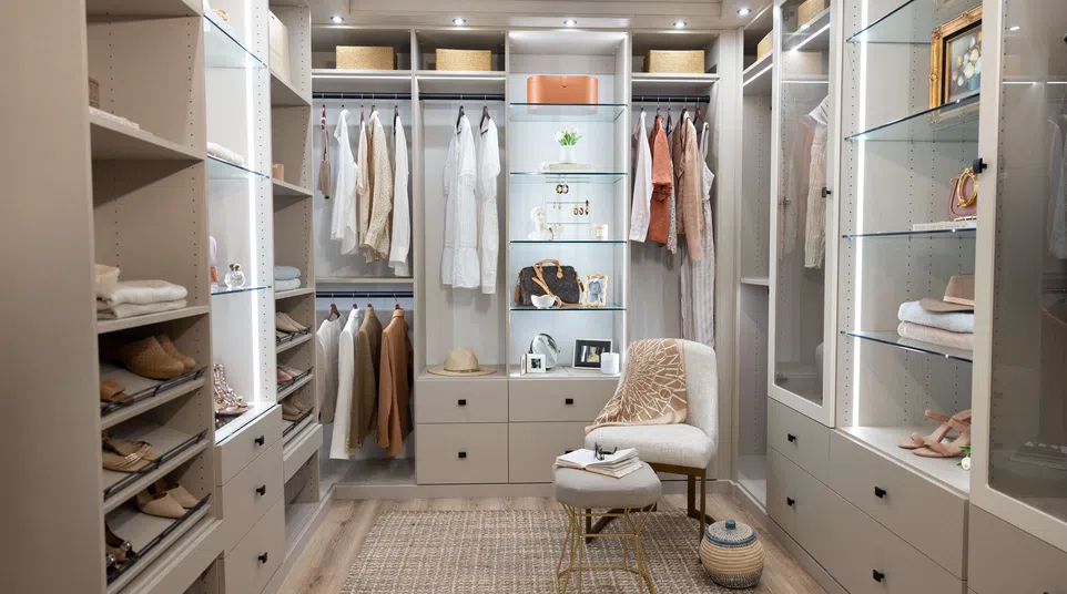Modern walk-in closet with neutral-toned clothing, glass shelves, padded chair, and woven rug on wood flooring.