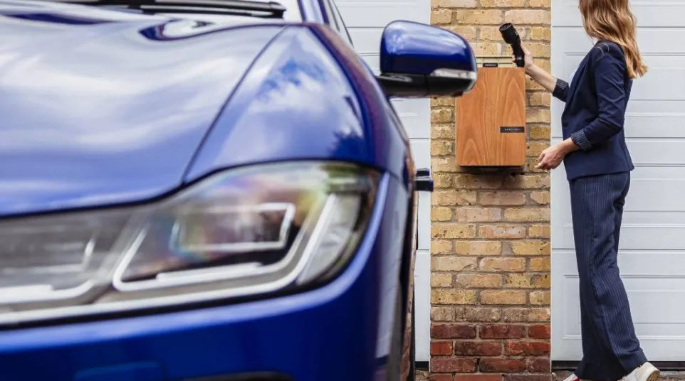 Woman charging a blue electric car using a wall-mounted home charger on a brick wall.
