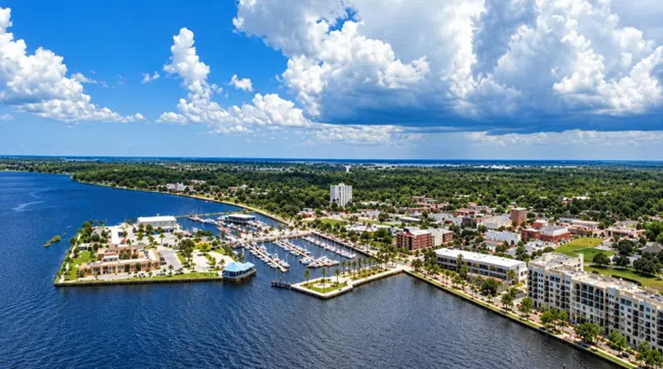 Aerial view of a coastal city with marina, residential buildings, greenery, and cloudy blue sky.