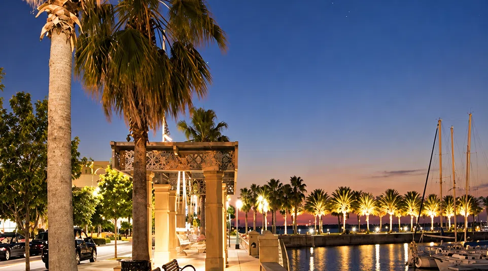 Evening waterfront promenade with palm trees, benches, sailboats, and lit walkway under a twilight sky.