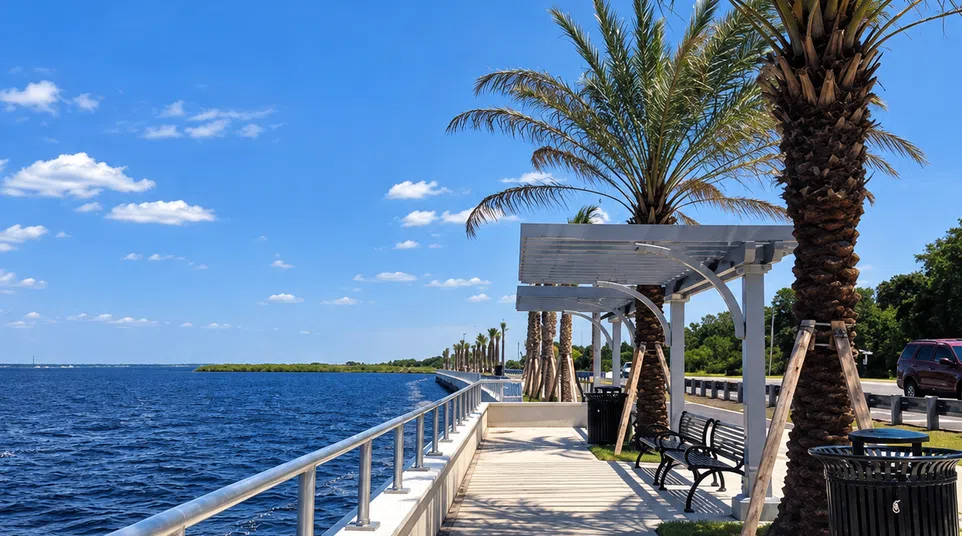 Waterfront promenade with palm trees, benches, metal railings, and a clear blue sky on a sunny day.