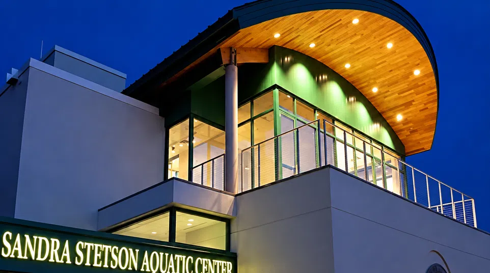 Modern Sandra Stetson Aquatic Center building illuminated at dusk with curved wooden roof and glass walls.