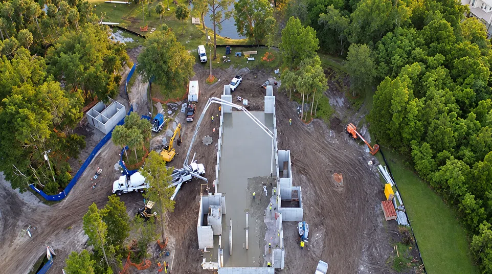 Aerial view of residential construction site near a lake with concrete foundation and surrounding trees.