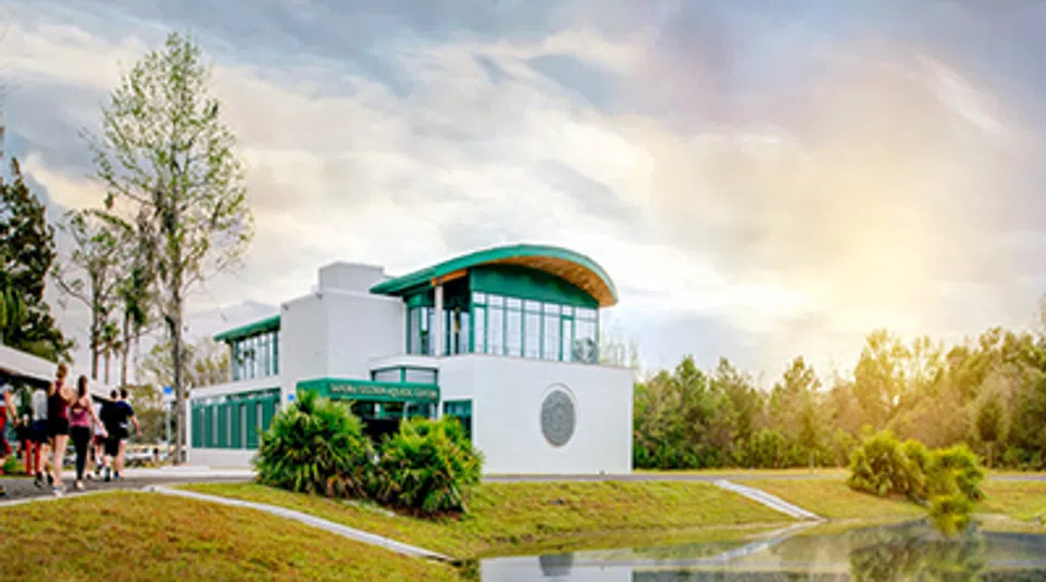 Modern white building with green roof by a reflective pond and people walking outdoors on a cloudy day.