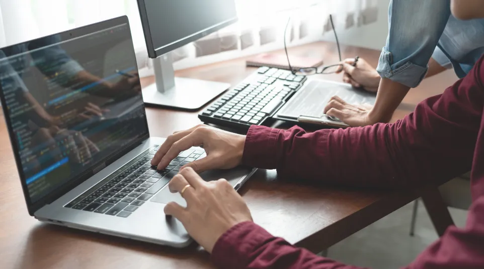 Person typing on a laptop with code on screen, another person reviewing data on a desk nearby.