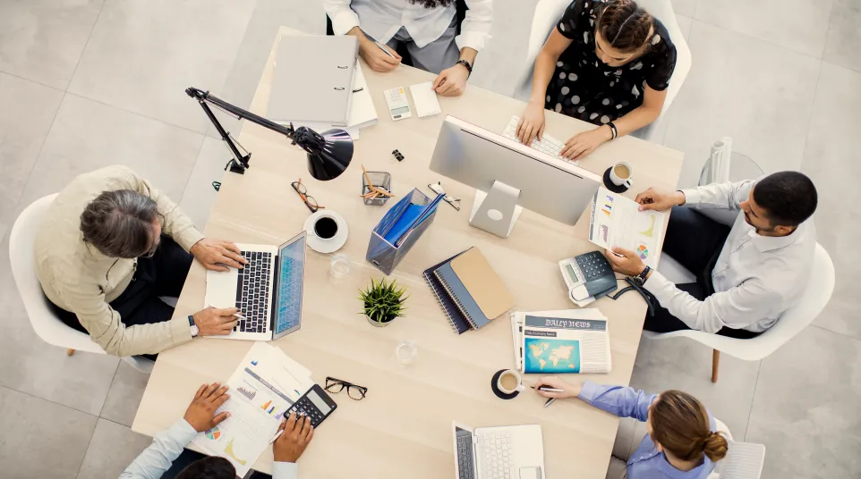 Top view of a diverse team collaborating around a modern office desk with laptops and documents.