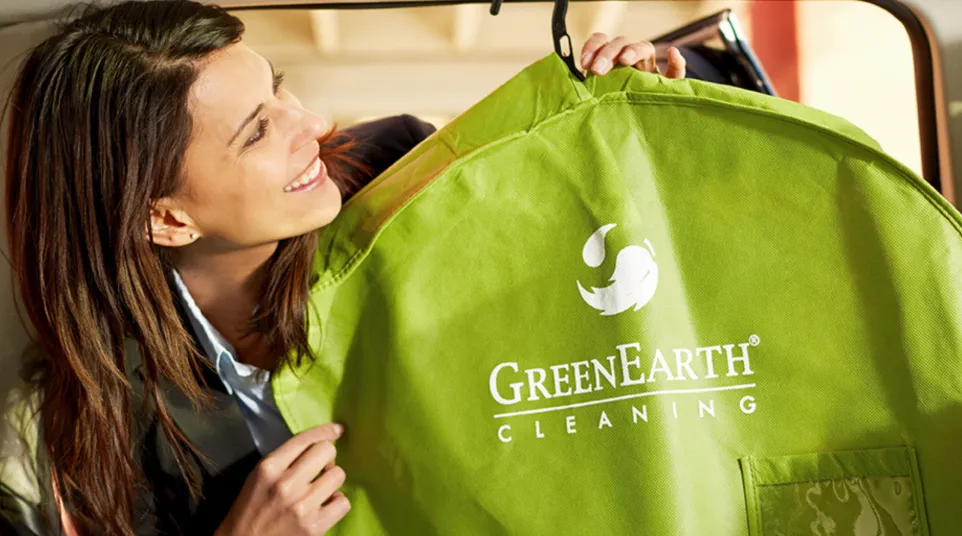 Woman holding a green garment bag with GreenEarth Cleaning logo inside a car, smiling and looking at bag