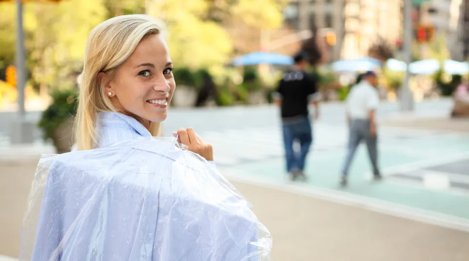 Smiling blonde woman carrying freshly dry-cleaned clothes in a plastic cover on city sidewalk.