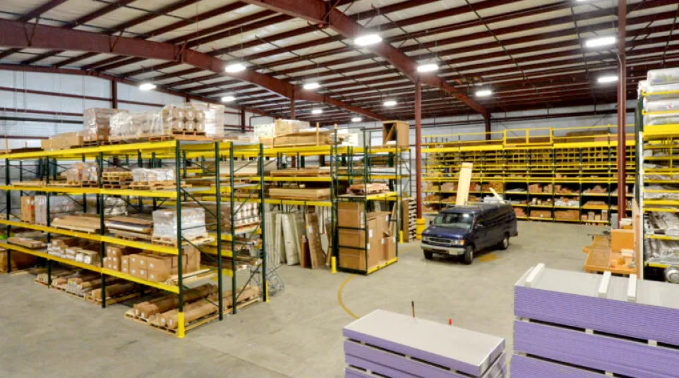 Spacious warehouse with tall shelves stocked with building materials and a black van parked inside under bright lighting.