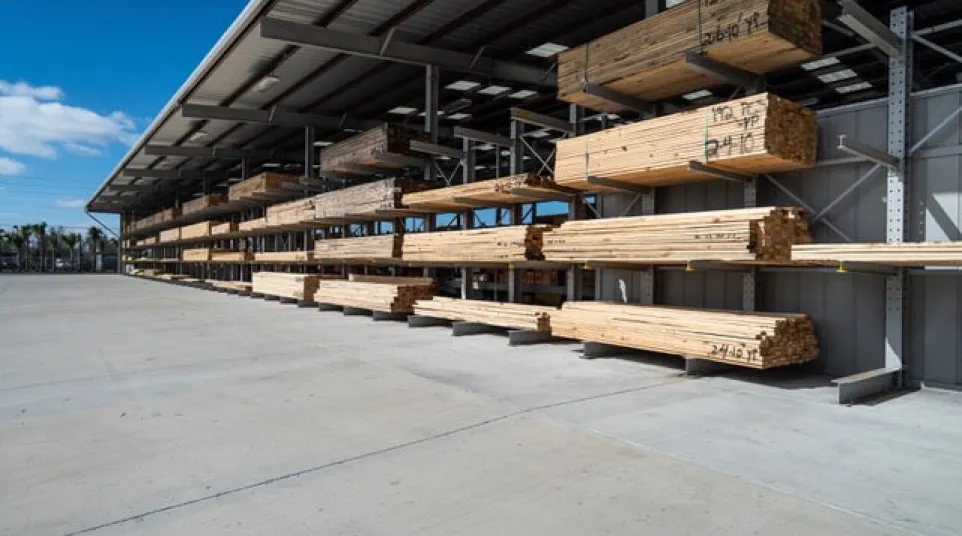 Stacks of lumber organized on metal racks under a large roof inside a spacious outdoor lumber yard.