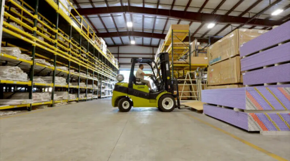 Worker operating a forklift in a warehouse moving stacked drywall sheets alongside storage racks.
