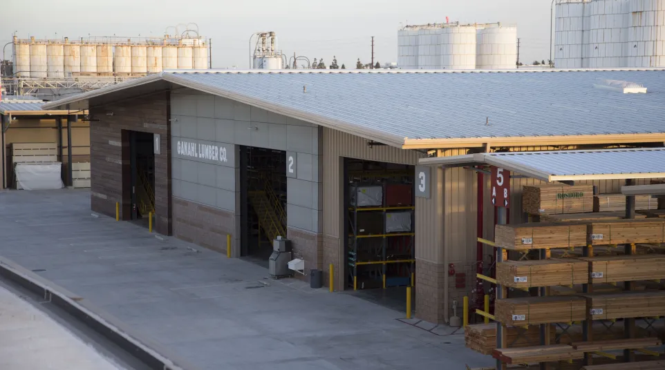 Ganahl Lumber Co. warehouse with stacked lumber and industrial tanks in the background under clear sky
