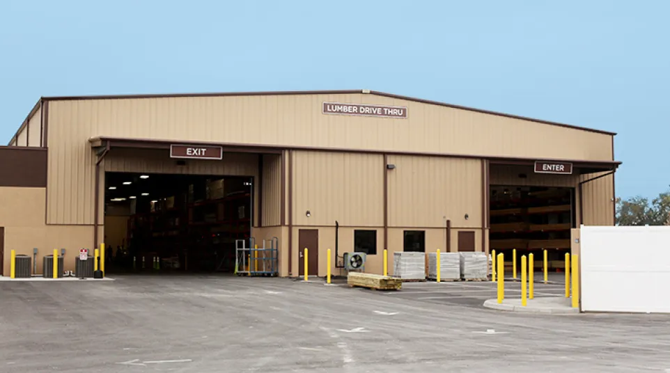 Exterior view of a lumber drive-thru with designated enter and exit bays and stacks of wood inside.
