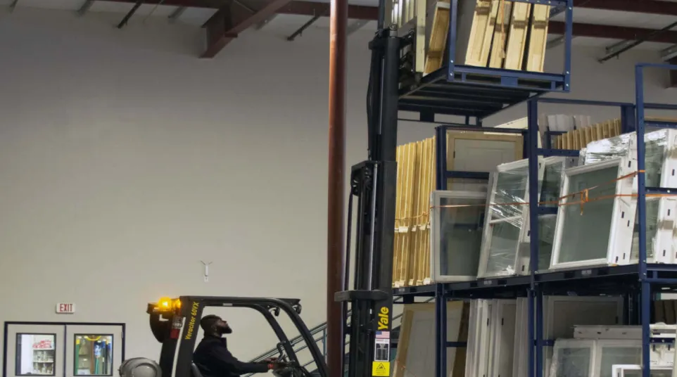 Man operating yellow Yale forklift lifting wooden frames in a warehouse with metal shelving and packaged windows.