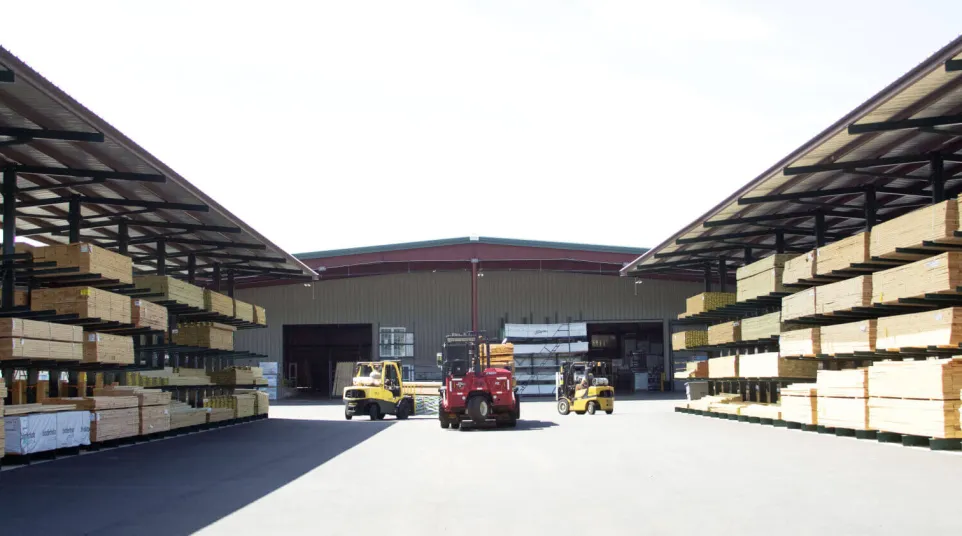 Industrial lumberyard with stacked wood, forklifts moving, and storage warehouses under bright sunlight.