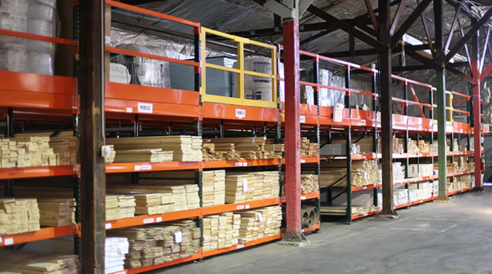 Warehouse shelves stocked with stacked wooden planks and materials under a high ceiling with metal beams.