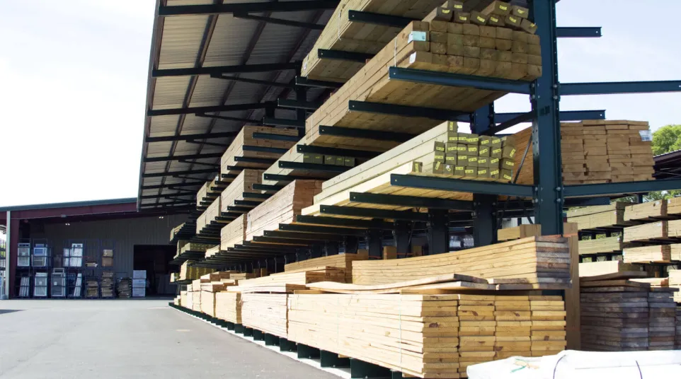 Stacks of various sizes of wooden planks stored on metal racks in an outdoor lumberyard with a partially covered roof.