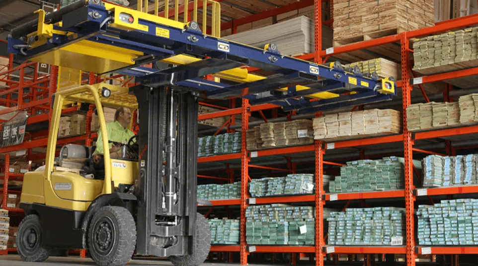 Forklift operator moving stacked materials in a large warehouse with orange metal shelving and wooden pallets.