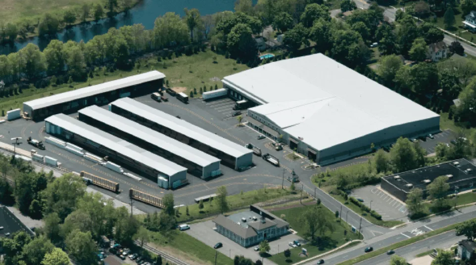 Aerial view of a large warehouse complex with multiple loading docks and parked trailers surrounded by trees and roads.