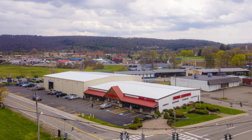 Aerial view of a commercial hardware and lumber store with parking lot and surrounding businesses under cloudy sky.