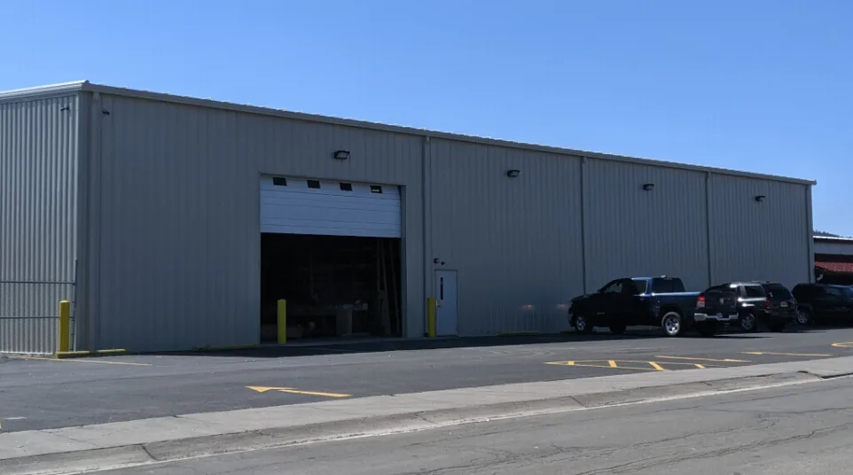 Gray industrial warehouse with open garage door and parked vehicles under clear blue sky.