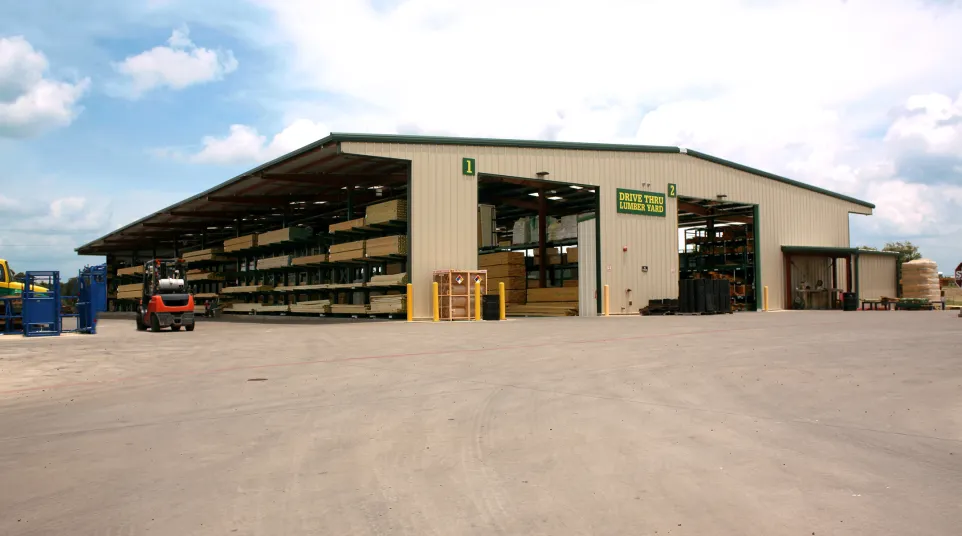 Large lumber yard building with stacked wood and forklift under partly cloudy sky.