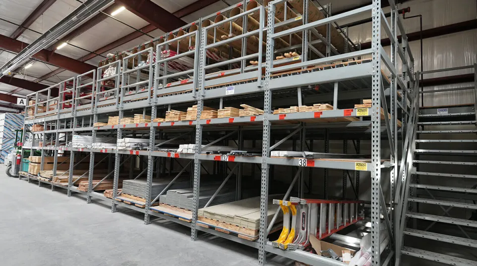 Warehouse shelving stocked with lumber, building materials, and tools under metal mezzanine level
